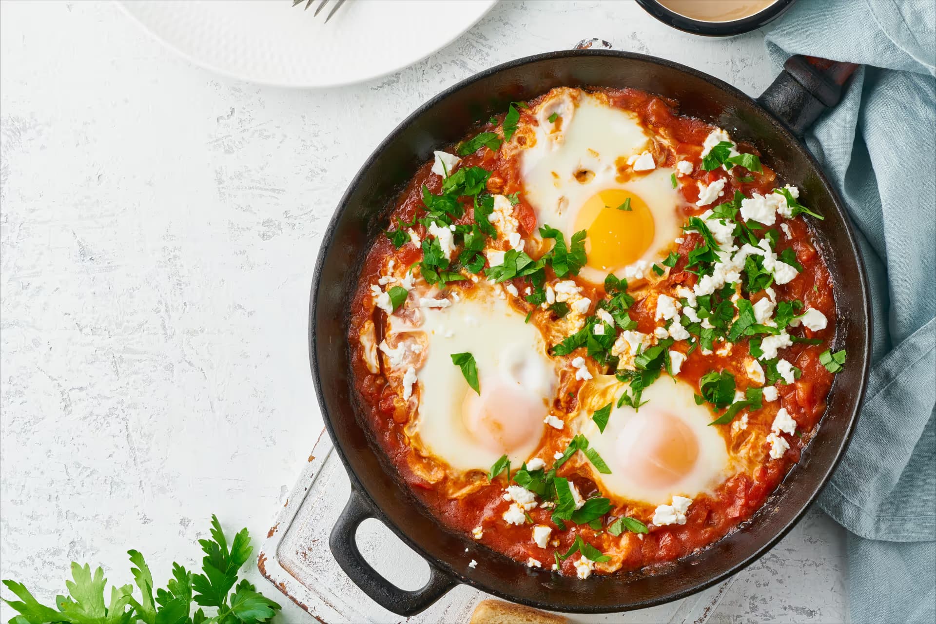 Shakshuka cu ouă, roșii, brânză feta și verdeață proaspătă, preparată în tigaie.